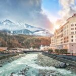 View of the houses, hotels, shops, the Mzymta River and the snowy peaks of the mountains on Rosa Khutor in Sochi on a sunny day.